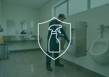 Technicien de maintenance en uniforme bleu et lunettes de sécurité brossant un urinoir collectif rincé à grande eau dans des sanitaires d'entreprise.